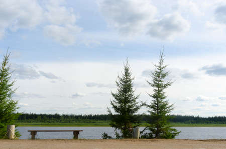 The road in the Northern Yakut village of ulus Suntar goes along the cliff above the vilyu river with small fir trees, a bench for rest with urns under the clouds on a summer day.の写真素材