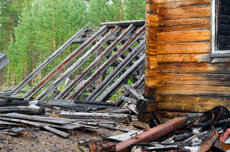 A corner of a burned-out wooden house with a window and a greenhouse roof next to yellow wood and black charred logs in the forest after the fire.の写真素材