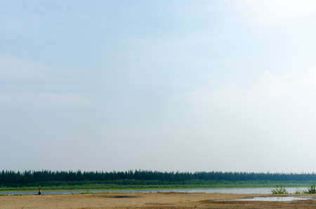 Girl traveler resting on the edge of the old pier on the banks of the river vilyu with spruce forests and the tundra of Yakutia in the Far North of Russia looking at the phone on a warm day.の写真素材