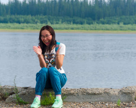 Joyful Yakut Asian young girl sitting on the stone Bank of the vilyu river and the tundra Northern forest in the village of Ulus Suntar clapping admiringly.の写真素材