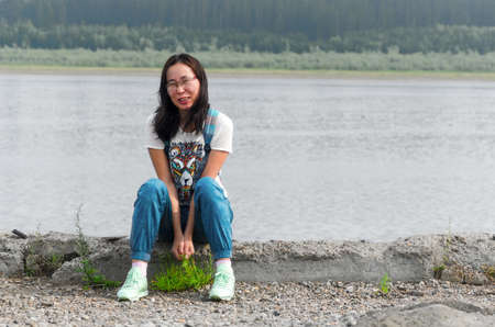 The girl traveler sits on the edge of the old pier on the Bank of the river vilyu with spruce forests and the tundra of Yakutia in the Far North of Russia and smiles with her hands folded between her legs.の写真素材