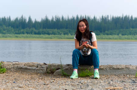 Cheerful sports Yakut Asian young girl sitting on the stone Bank of the river Viluy in the Northern forest puts his hands in the castle and smiles.の写真素材