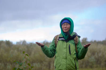 Yakut Asian girl tourist in a jacket hat and hood and with a backpack is meditating, closing his eyes, under the hail and rain raising his hands against the background of the wild taiga of the North in the wind.の写真素材