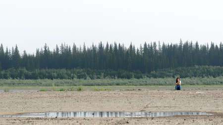 Girl traveler resting on the edge of the old pier on the banks of the river vilyu with spruce forests and the tundra of Yakutia in the Far North of Russia looking at the phone on a warm day by a puddle.の写真素材