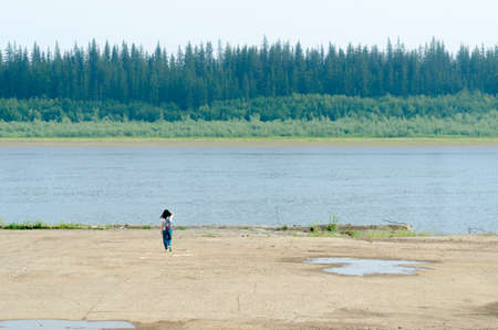 Girl traveler goes to the edge of the pier on the shore, holding her hair to the river vilyu with spruce forests and the tundra of Yakutia in the Far North of Russia.の写真素材