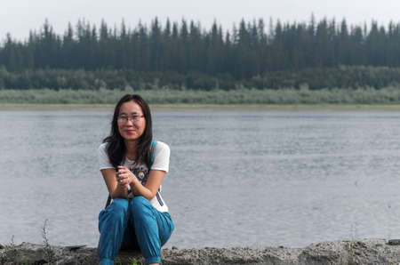 A thoughtful girl-traveler sits on the edge of an old pier on the Viluy river with the forests of Yakutia in the Far North of Russia,with a smile on her face.の写真素材