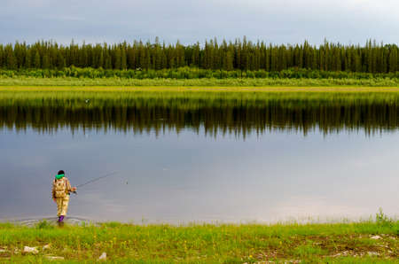 Yakut girl tourist fisherman with a backpack in boots comes into the Northern river vilyu with a spinning in his hand in the background of the wild forest taiga.の写真素材