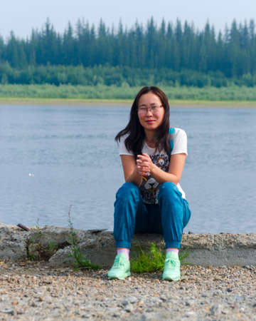 Brooding Yakut Asian young girl sitting on the stony shore, and smiling with folded hands in the background of the Vilyui river and tundra.の写真素材