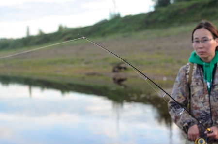Yakut Asian girl-the tourist-fisherman with glasses carefully monitor the tip of the rope spinning in a river on the background of the taiga and trees of the forest.の写真素材