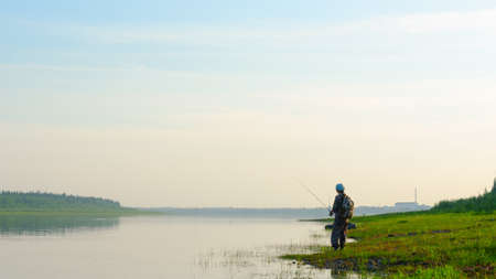 Yakut Asian girl tourist fisherman with a backpack and a cap to fish in the river vilyu in the haze at sunset in the wild North of Russia on the background of the village Suntar.の写真素材