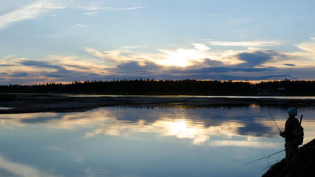 Yakut girl tourist with a backpack angler fishing and throws spinning on the wild Northern river in the tundra spruce forests of Russia holding a second fishing rod between his legs bright evening.の写真素材