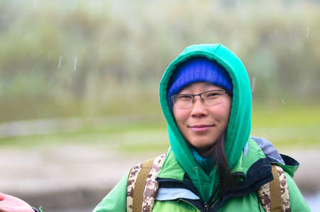 Yakut Asian girl tourist in hat and hood smiling with glasses on the background of snowy weather and green grass in the forest near the river in the North.の写真素材