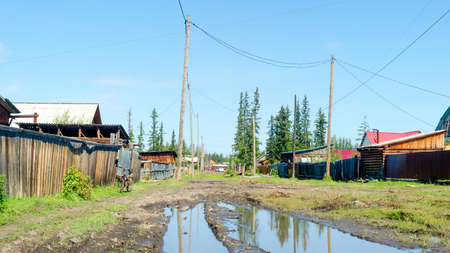 Bright day in the village of ulus Suntar in Yakutia on a residential street with a large puddle and a car in the distance among the wooden houses.の写真素材
