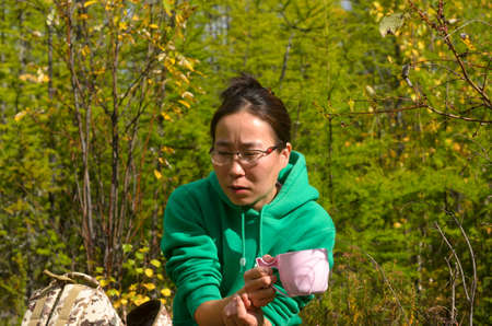 Thoughtful Yakut Asian girl tourist with glasses sitting with a Cup of tea next to a backpack in the wild forest of Yakutia.の写真素材
