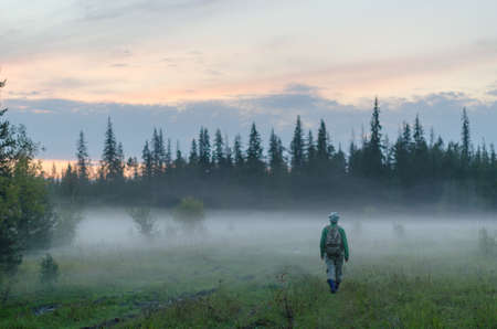 A tourist girl with a backpack goes into the fog in the forest in the evening at sunset in the wild taiga of Northern Yakutia in Russia.の写真素材