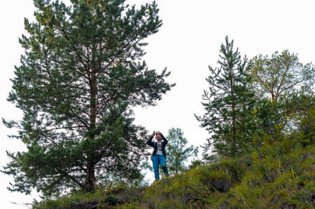 Yakut Asian girl tourist takes pictures on the phone view from the mountain in the spruce forest of Yakutia.の写真素材