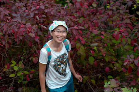 A cheerful smiling Asian tourist girl in glasses stands in the red autumn leaves of the trees below and looks up.の写真素材