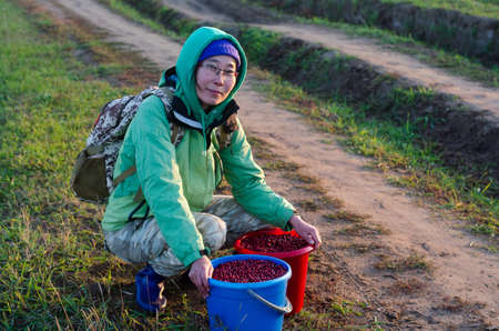 Yakut Asian tourist girl with backpack, glasses and cap with hood sits near the road with two buckets full of collected wild cranberries in the North.の写真素材