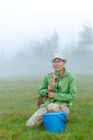 Yakut Asian girl in a cap and glasses sitting smiling and holding the strap of a backpack at the bucket before picking wild berries in the fog white behind.の写真素材
