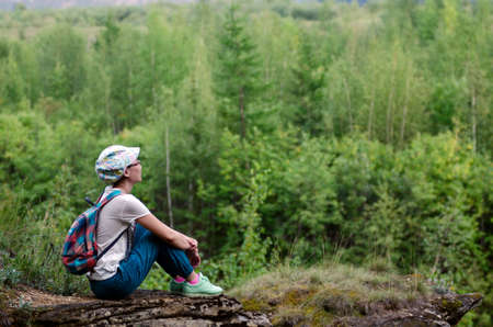 Yakut Asian girl tourist with backpack and cap sitting on the edge of the mountain in the wild tundra of Yakutia hugging his legs resting and looking into the distance.の写真素材