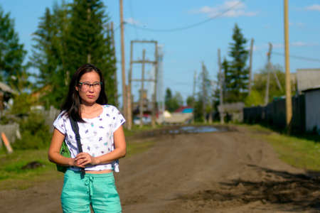 Fashionable modern Yakut girl with glasses and a handbag is thoughtful on the village road among the trees of fir trees and fences in Northern Yakutia.の写真素材