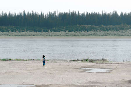 Girl traveler with backpack goes to the edge of the concrete sidewalk touching the hair on the Bank of the old pier North of the river vilyu with spruce forests and the tundra of Yakutia on the other side.の写真素材