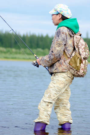 Yakut girl tourist fisherman fishing spinning standing in the water in boots stranded rifts of the Northern river vilyu and tundra spruce forest.の写真素材