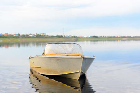 Old metal boat is in calm water with reflection of clouds, was chained against the background of the houses of the Yakut of the Northern villages in the forest Suntar.の写真素材