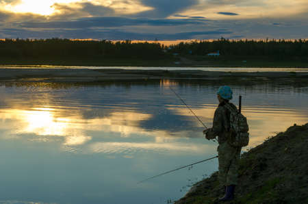 Girl fisherman tourist with a backpack holding one rod in his legs throws a second spinning on the background of a bright sunset in the wild taiga of Yakutia.の写真素材