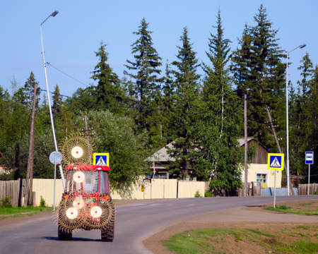 A tractor with a device for collecting hay rides on the road among the houses of the Northern village of Suntar in Yakutia against the background of large fir trees in the forest.

の写真素材