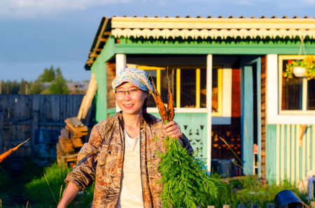 Yakut joyful Asian girl with glasses and a cap and carries a bundle of freshly harvested carrots going on the residential area North of the Yakut village.の写真素材