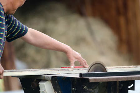 A man neatly saws a Board on a circular saw against the background of a shed with wood.の写真素材