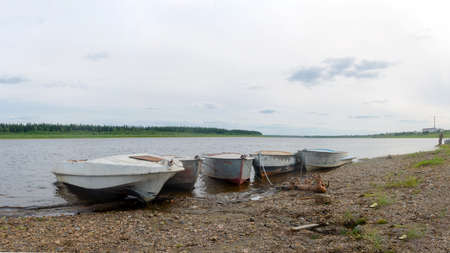 Five old metal boats are tethered near the shore against the background of a girl fisherman and the village of Northern Yakutia in the taiga Suntar.の写真素材