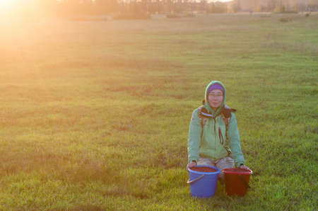 Yakut Asian girl in a hood and hat, and wearing glasses sitting on a green field on the background of spruce forest taiga in the sunset about two collected buckets of wild cranberries in the North.の写真素材