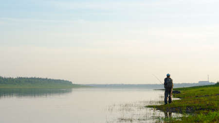 Yakut Asian girl-tourist fisherman with a backpack and a hat to fish with a spinning in the river Vilyuy in the haze at sunset in the wild North of Russia on the background of the village Suntar.の写真素材