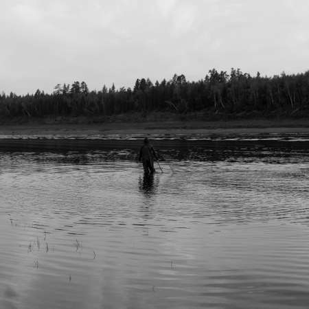 Black and white photo of a Lonely man, a Yakut fisherman in wading boots wearily goes to the net for wildlife in the river Vilyuy in the bright rays of the sun catching the local fish chugunok at sunset .の写真素材