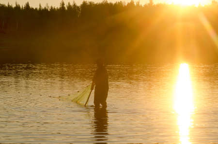 A lonely man, a Yakut fisherman in wading boots wearily goes to the net for wildlife in the river Vilyuy in the bright rays of the sun catching the local fish chugunok at sunset.の写真素材