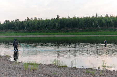 Two men of the Yakuts in the background gulls are in wading boots with fish net along the shores of the wild North of Vilyuy river in a forest traditionally catching local fish of tugun.の写真素材