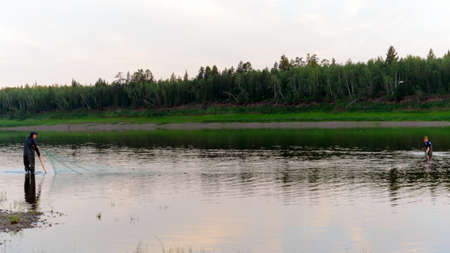 Two Yakut men turn around walking in waders with a fishing net along the banks of the wild North of the Viluy river in the forest, traditionally catching local tugun fish.の写真素材