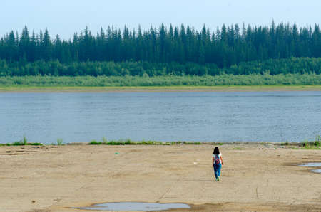 Girl tourist lonely goes to a concrete cliff on the shore of the old pier at the Northern river vilyu with spruce tundra forests of Yakutia on the other side.の写真素材