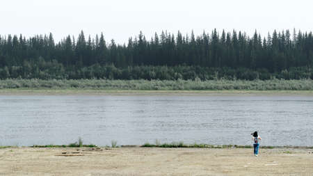 The girl-traveler goes to the edge of the pier on the shore, holding the straps of a backpack to the North of the vilyu with spruce forests and the tundra of Yakutia on the other side of the shore.の写真素材