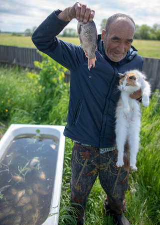 A village old man holds a carp in one hand and a cat looking at food in the other against the background of a bath with fish behind the fence.の写真素材