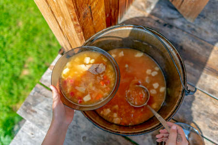 Hands girls cook outdoors lays out soup, lunch with meat and vegetables and potatoes, borscht from a large pot on the wooden floor, in the house next to the grass field, in transparent plates.の写真素材