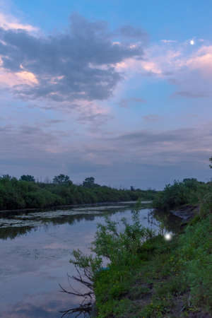 The reflection of the moon and clouds sky sunset at dusk in the water of the river near the banks with green grass and trees.の写真素材