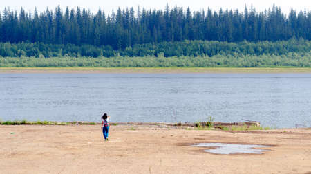 A tourist girl with a backpack touching her hair, lonely goes to the concrete cliff of the shore on the old pier at the Northern river vilyu with spruce tundra of the forest of Yakutia on the other side.の写真素材