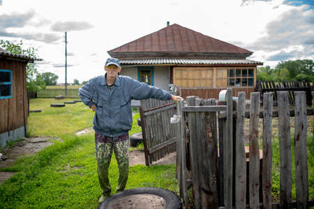 An old man in glasses and a cap in pants stained with cut grass stands at the village house holding on to the fence.の写真素材