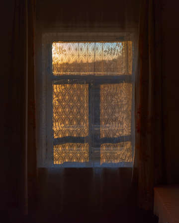A dark, rustic room with a corner table and a window behind a curtain with the setting sun at sunset.の写真素材