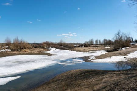 Spring Bay of the river with clear water and ice and snow on the sandy shore in the afternoon under the blue sky.の写真素材
