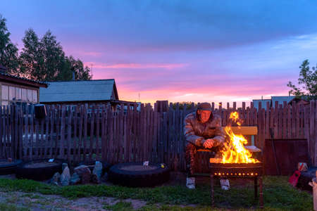 An old lonely man sits sadly on a bench at a village house behind a fence looking into the fire of a barbecue fire at sunset in the evening.の写真素材
