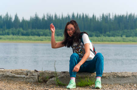 Joyful Yakut Asian young girl sitting on the stone Bank of the river Viluy in the Northern forest throws a stone forward.の写真素材